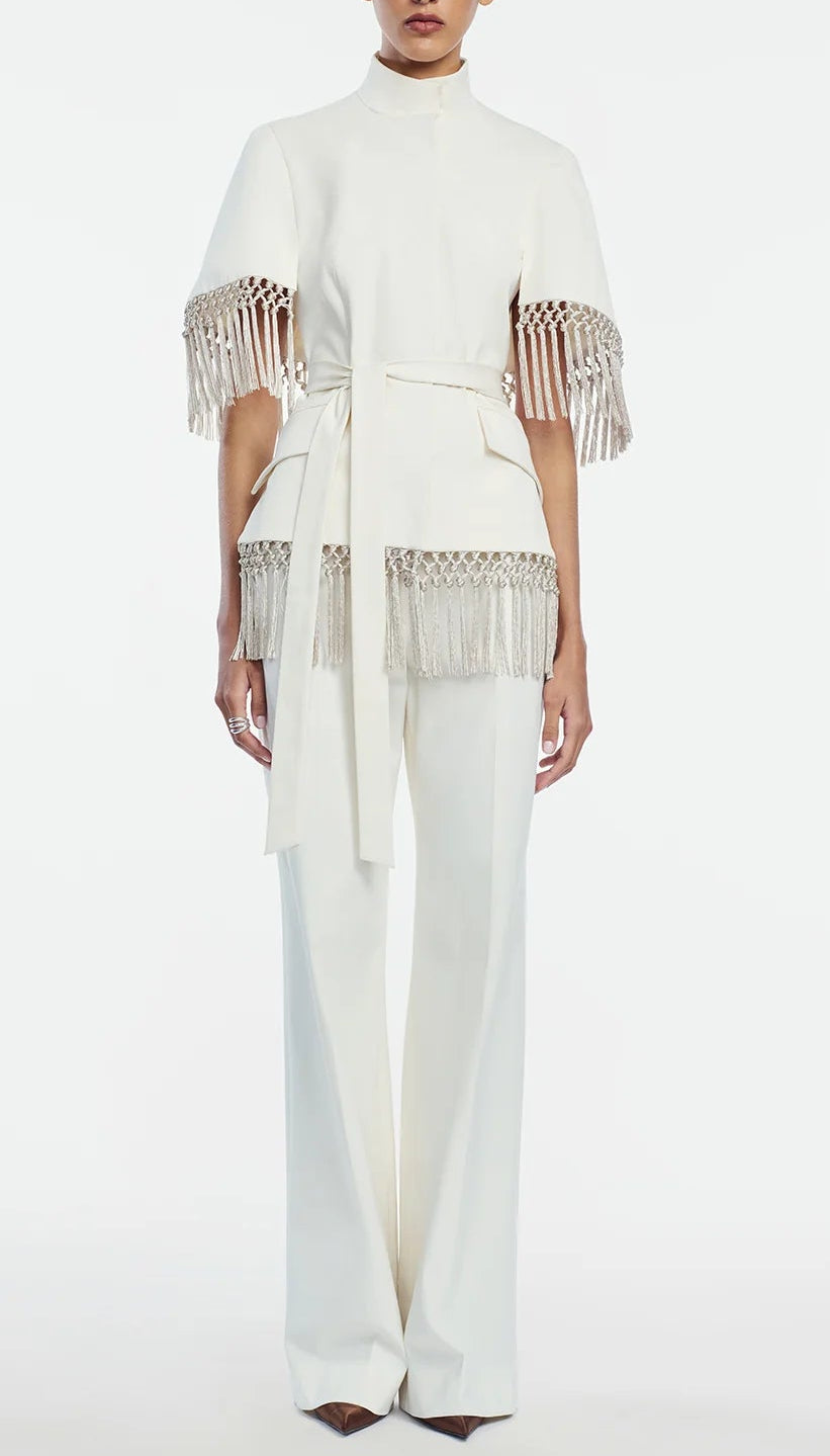 A person wearing the Lela Rose Stretch Cotton Molly Pant, styled with a white tailored pantsuit crafted from stretch cotton, featuring a belted top with fringe details on the sleeves and waist and paired with high-rise straight leg pants for a modern, polished look.