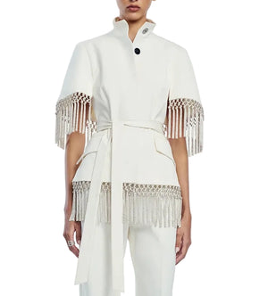 A person wearing the Lela Rose Stretch Cotton Fringed Cape—a white, short-sleeved, belted jacket with decorative beaded fringe detailing on the sleeves and hem, inspired by western dressing—paired with matching white pants.