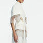 A person wearing the Lela Rose Stretch Cotton Fringed Cape—a white, belted jacket featuring metallic fringe detailing on the sleeves and hem that blends elements of western dressing—stands against a plain background.
