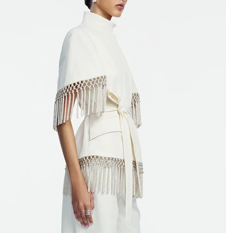A person wearing the Lela Rose Stretch Cotton Fringed Cape—a white, belted jacket featuring metallic fringe detailing on the sleeves and hem that blends elements of western dressing—stands against a plain background.