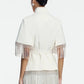 A woman facing away, wearing the Lela Rose Stretch Cotton Fringed Cape—a white, belted jacket with short sleeves and fringe detailing at the sleeves and hem, styled in a contemporary Western fashion—paired with white pants.