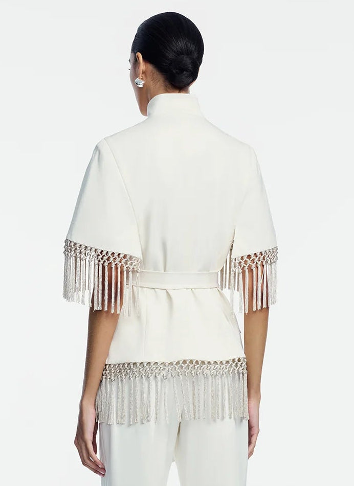A woman facing away, wearing the Lela Rose Stretch Cotton Fringed Cape—a white, belted jacket with short sleeves and fringe detailing at the sleeves and hem, styled in a contemporary Western fashion—paired with white pants.