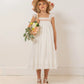 A young girl wears the Bonpoint Girls' Laora Dress, smocked and hand-embroidered, as she holds a bouquet against a white wall with a light wooden floor.