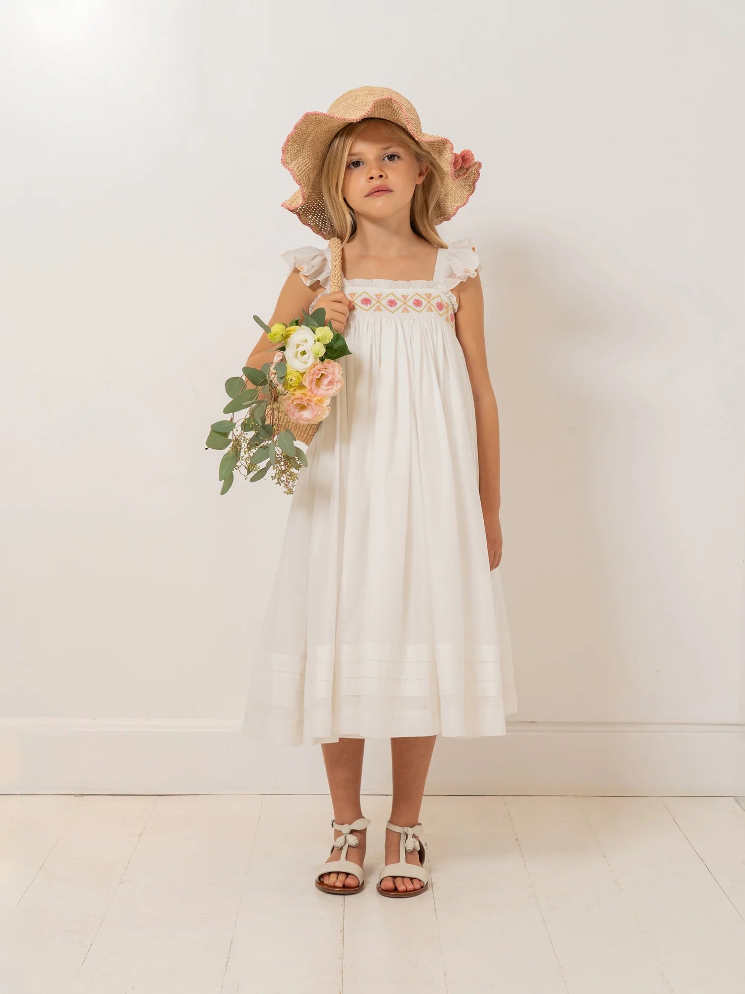 A young girl wears the Bonpoint Girls' Laora Dress, smocked and hand-embroidered, as she holds a bouquet against a white wall with a light wooden floor.