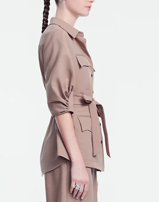 A woman stands in profile against a plain light background, wearing the Lela Rose Melange Crepe Camp Shirt in taupe melange, featuring chest pockets and a belt.
