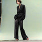 A person stands against a green backdrop wearing a loose black blazer, TWP Adieu for Now Sequin Pant, and red strappy sandals.