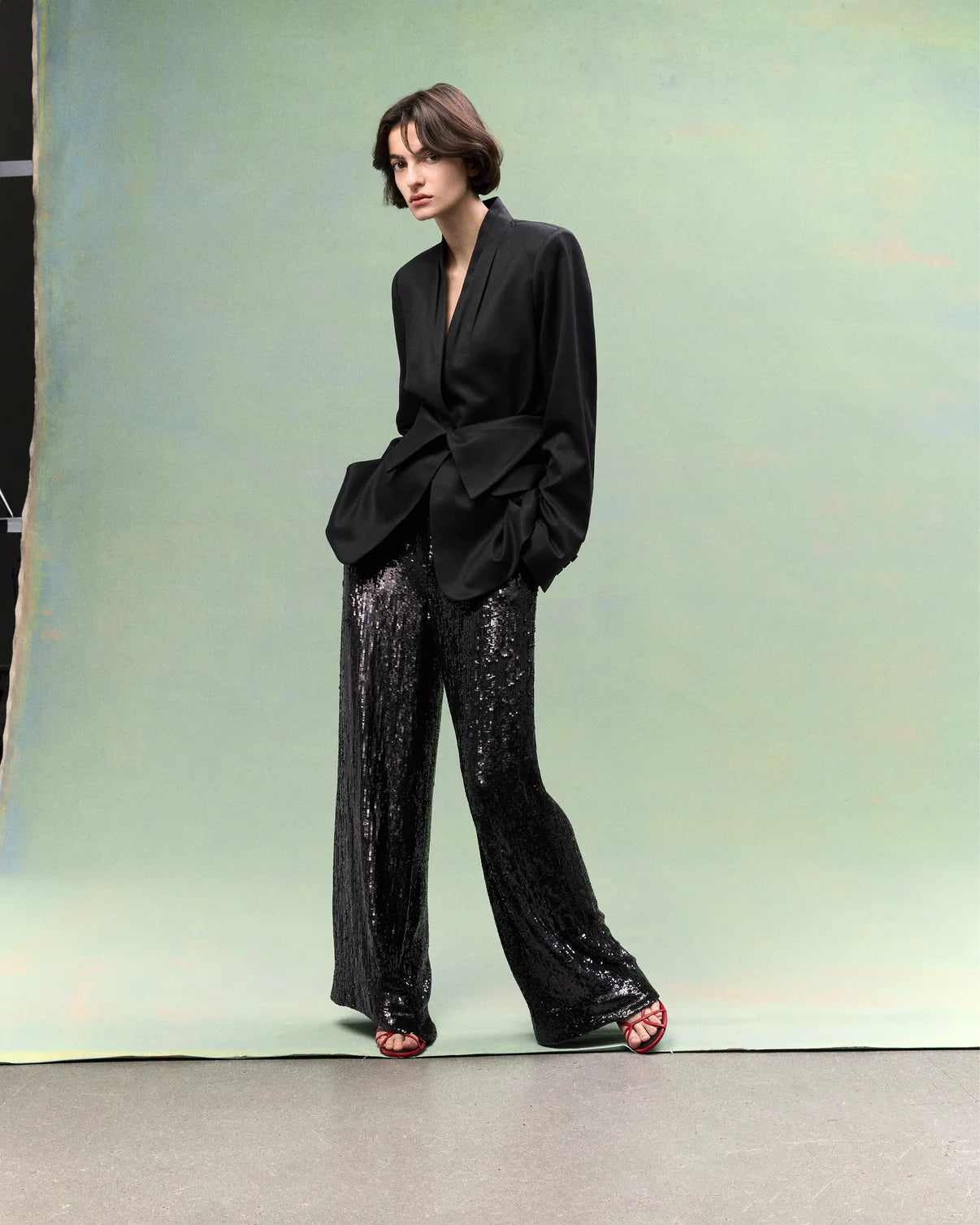 A person stands against a green backdrop wearing a loose black blazer, TWP Adieu for Now Sequin Pant, and red strappy sandals.