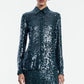 A person wearing the Lela Rose Sequin Lucy Shirt—a long-sleeve, dark green sequin top—paired with a matching skirt, stands against a plain white background. The Sequin Lucy Shirt from Lela Rose is a bold statement piece that effortlessly elevates the entire look.