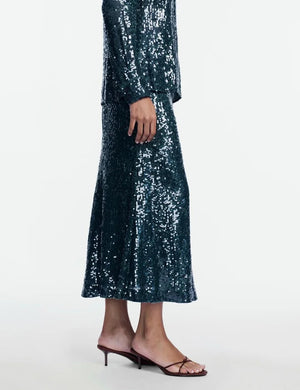 A person is dressed in the Lela Rose Sequin Skirt paired with a matching top, featuring shimmering sequins and styled with strappy heeled sandals, standing against a plain white background.