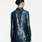 A person with dark hair in a low bun is shown from behind, wearing a statement piece: the Lela Rose Sequin Lucy Shirt—a long-sleeved, dark green sequin top—paired with a matching skirt against a plain white background.