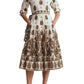 A woman wearing the Cara Cara Lolita Dress—a long-sleeve, collared midi dress featuring a white and brown paisley border print with pockets—paired with brown heels, stands against a white background.