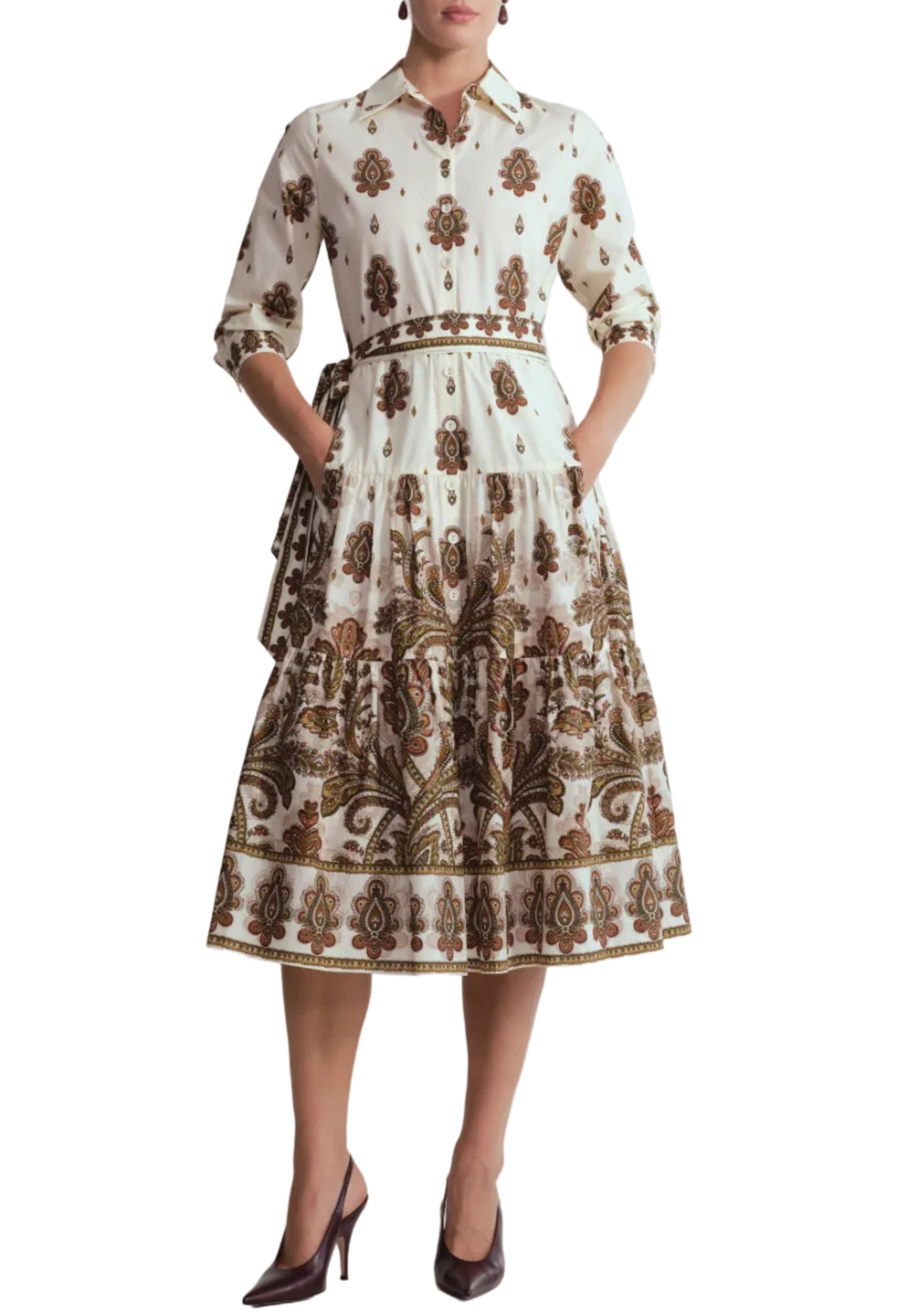 A woman wearing the Cara Cara Lolita Dress—a long-sleeve, collared midi dress featuring a white and brown paisley border print with pockets—paired with brown heels, stands against a white background.