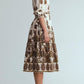 A woman stands in profile against a plain background, wearing the Cara Cara Lolita Dress—a white, long-sleeve, collared midi dress with a paisley border print and a belt—paired with dark brown slingback heels.