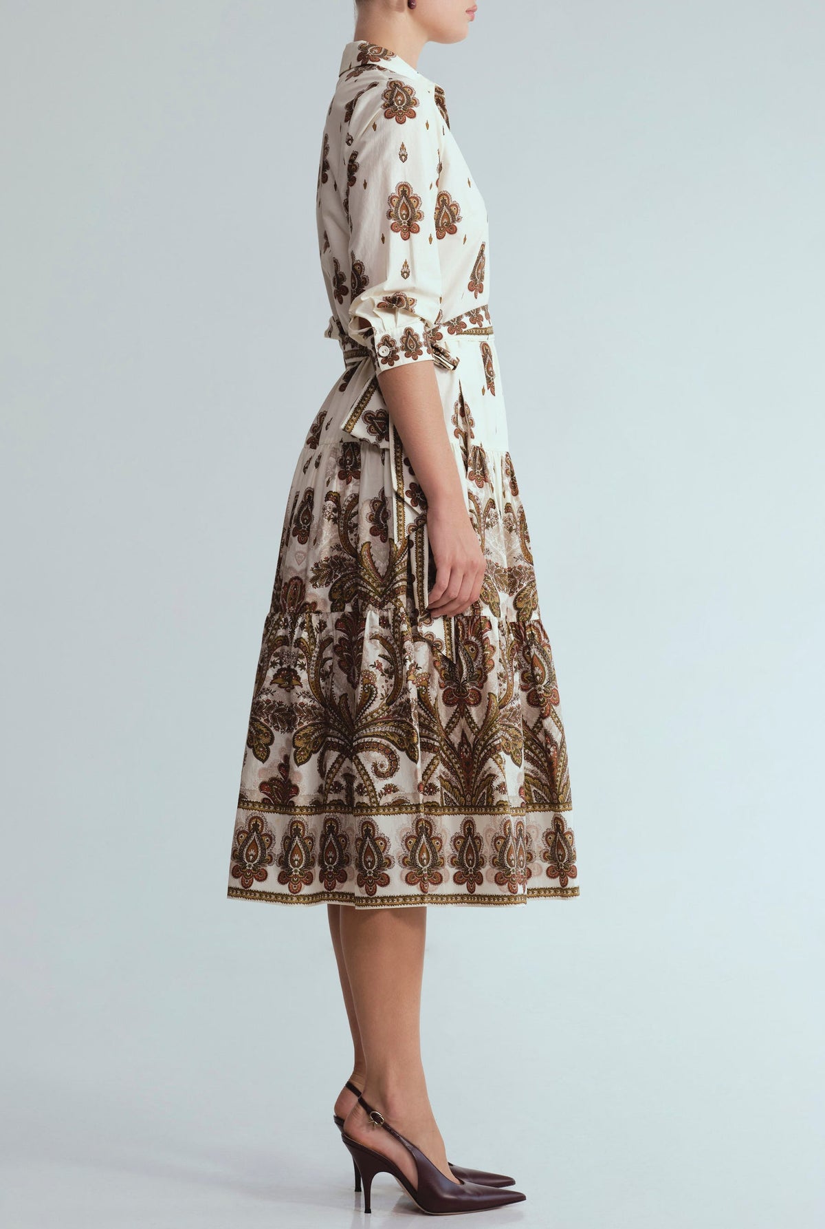A woman stands in profile against a plain background, wearing the Cara Cara Lolita Dress—a white, long-sleeve, collared midi dress with a paisley border print and a belt—paired with dark brown slingback heels.