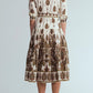 A person stands facing away against a plain background, wearing the Cara Cara Lolita Dress—a collared, midi white dress with brown and gold patterns—paired with brown heels.