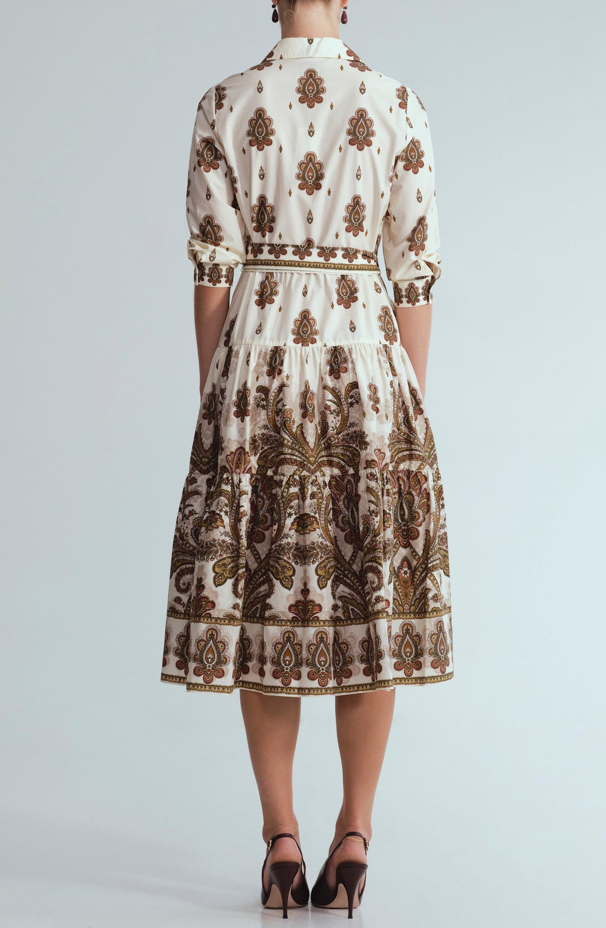 A person stands facing away against a plain background, wearing the Cara Cara Lolita Dress—a collared, midi white dress with brown and gold patterns—paired with brown heels.