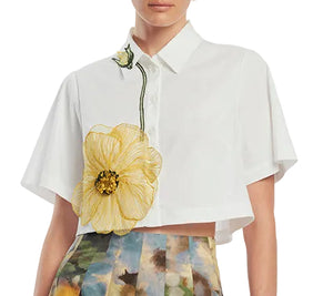 A woman wears the Lela Rose Peony Embroidered Cropped Shirt, a white cotton top with a large yellow flower detail, paired with a multicolored abstract-pattern skirt.