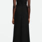 A person stands facing away, wearing the Lela Rose 3D Peony Embroidered Fluid Crepe Twisted Detail Dress—an ankle-length, sleeveless black dress with a scoop back—paired with beige heeled shoes against a plain white background.