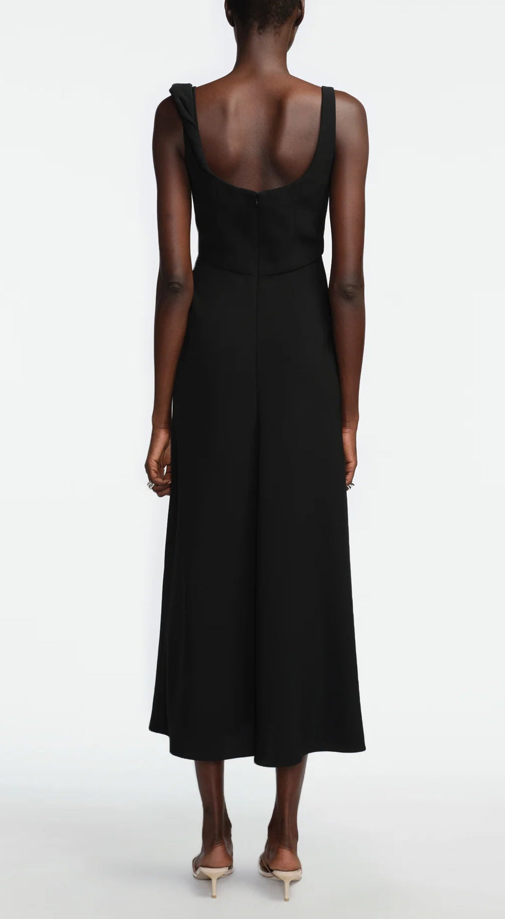 A person stands facing away, wearing the Lela Rose 3D Peony Embroidered Fluid Crepe Twisted Detail Dress—an ankle-length, sleeveless black dress with a scoop back—paired with beige heeled shoes against a plain white background.