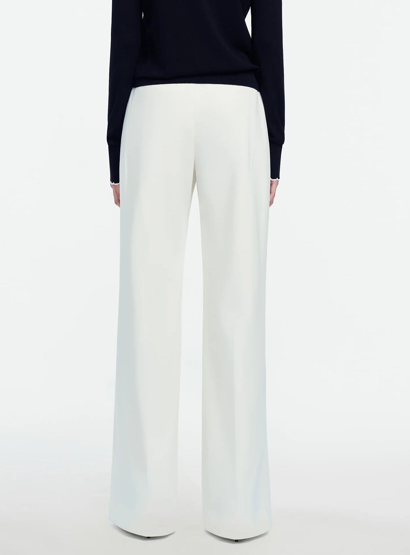 A person stands facing away against a plain white background, wearing a black long-sleeve top paired with the Lela Rose Stretch Cotton Molly Pant—high-waisted, wide-leg white pants featuring a high-rise silhouette.