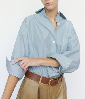 A person wears the TWP New Earl Shirt, an oversized light blue Italian cotton button-down with rolled sleeves, tucked into high-waisted tan trousers with a brown belt, standing against a plain white background.