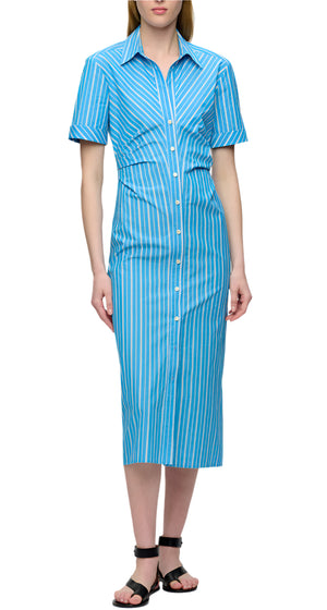 A woman wears the Lou Dress by Callas Milano, a blue and white vertical striped Italian cotton twill shirt dress with short sleeves and midi length, styled with black sandals against a white background.