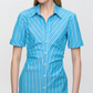 A person wearing the Lou Dress by Callas Milano, featuring blue and white stripes in Italian cotton twill, with short sleeves and a collar, stands against a plain background.