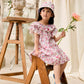 A young girl wearing the Louise Misha Girls’ Natiyana Dress, featuring floral cotton fabric and ruffled sleeves, sits on a wooden stepladder with a hat and holds a flower. A bust statue and another flower rest on the nearby table, while cream fabric drapes in the background.