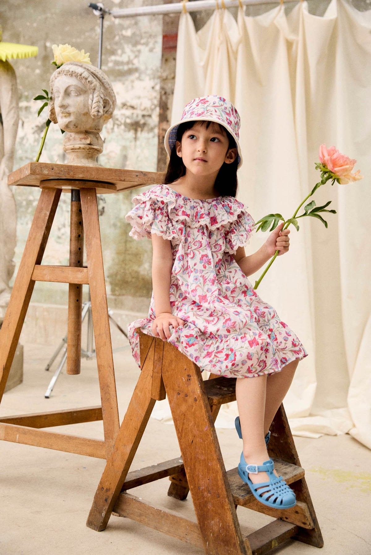 A young girl wearing the Louise Misha Girls’ Natiyana Dress, featuring floral cotton fabric and ruffled sleeves, sits on a wooden stepladder with a hat and holds a flower. A bust statue and another flower rest on the nearby table, while cream fabric drapes in the background.