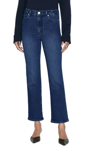 A person stands against a white background wearing Frame’s Le Sleek Straight dark blue high-rise straight-leg jeans, a dark long sleeve top, and black pointed-toe shoes.