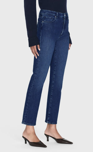 A person wearing Frame’s Le Sleek Straight high-rise, dark blue jeans, a long-sleeve navy top, and black low-heeled shoes stands against a plain light background.