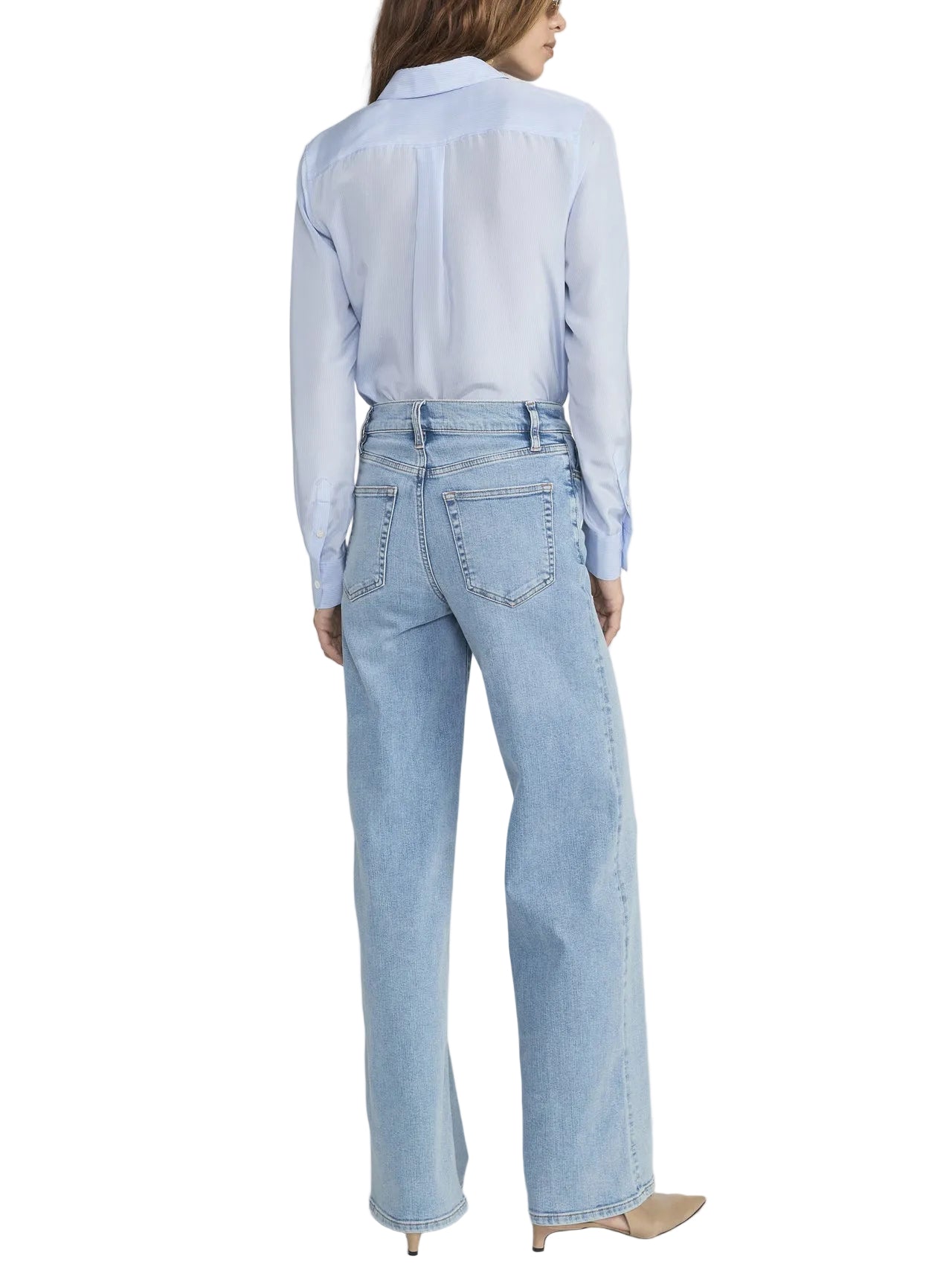 A person stands facing away in a light blue button-up shirt, Frame’s Le Slim Palazzo high rise light blue jeans crafted from sustainable cotton, and beige pointed shoes against a plain background.