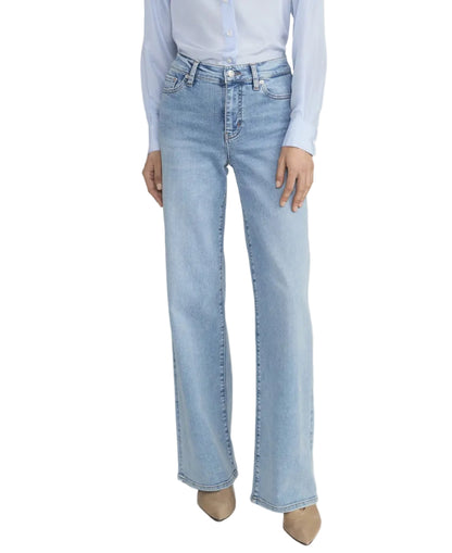 A woman stands against a plain background wearing Frame's Le Slim Palazzo high-rise, light blue jeans, a light blue button-up shirt, and beige pointed-toe shoes.