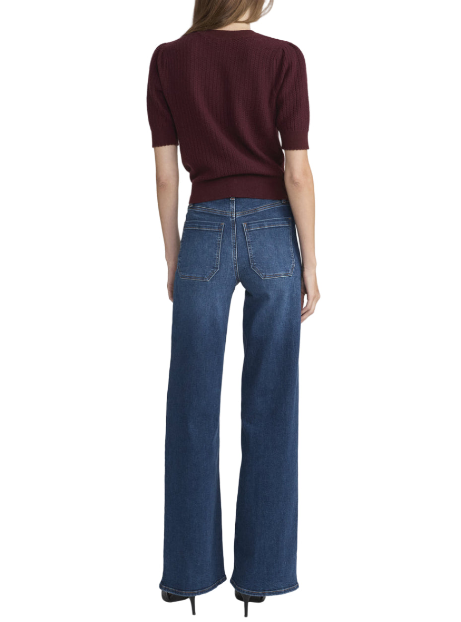 A woman stands facing away, wearing a maroon short-sleeve sweater and Frame’s Le Slim Palazzo Modernist Pocket high-rise sustainable cotton palazzo pants in blue with black heels against a plain white background.
