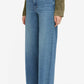 A person wearing Frame’s Le Slim Palazzo jeans in blue, a green jacket, and beige pointed-toe shoes, standing against a plain white background.
