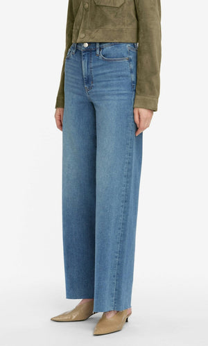 A person wearing Frame’s Le Slim Palazzo jeans in blue, a green jacket, and beige pointed-toe shoes, standing against a plain white background.