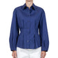 A person wears the Callas Milano Lucia Top, a long-sleeved, button-front blue shirt with a collar, paired with white pants against a plain background.