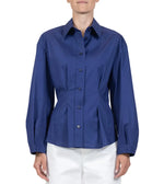 A person wears the Callas Milano Lucia Top, a long-sleeved, button-front blue shirt with a collar, paired with white pants against a plain background.