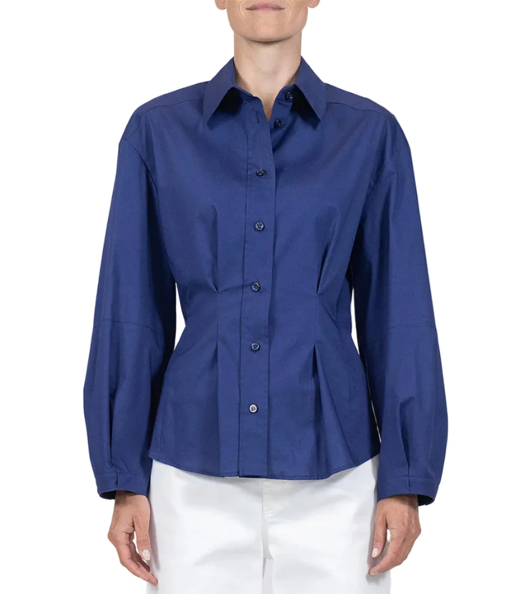 A person wears the Callas Milano Lucia Top, a long-sleeved, button-front blue shirt with a collar, paired with white pants against a plain background.