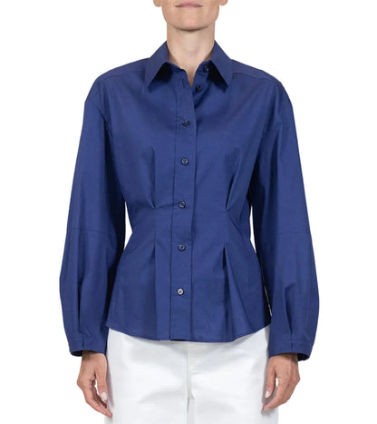 A person wears the Callas Milano Lucia Top, a long-sleeved, button-front blue shirt with a collar, paired with white pants against a plain background.