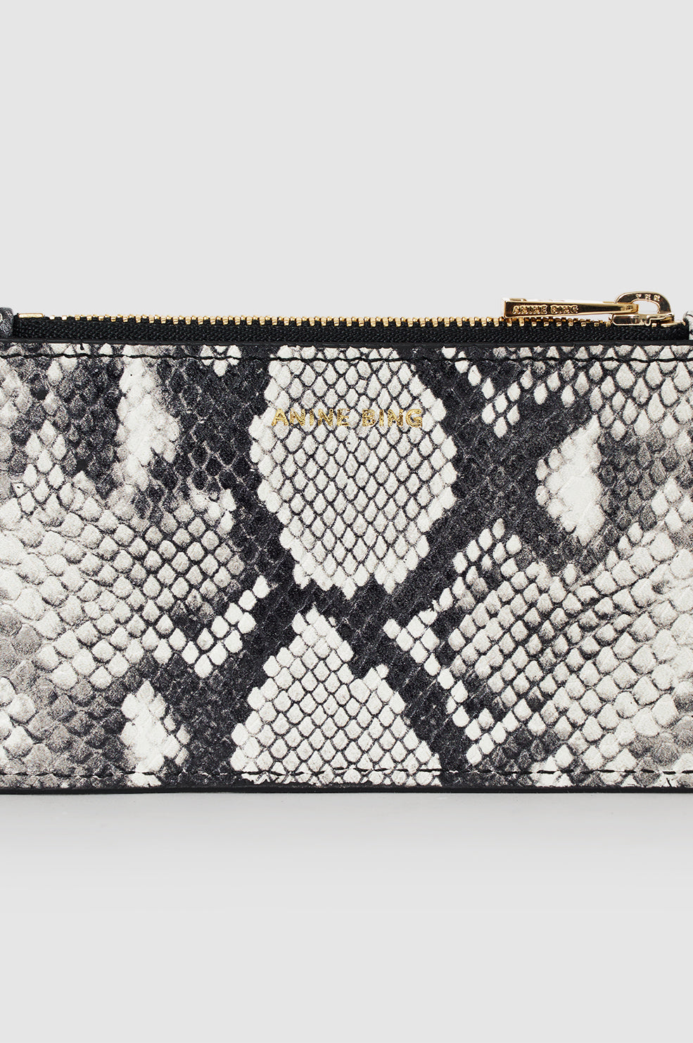 The Lucy Card Holder by Anine Bing is a compact rectangular wallet with a bold black and white python print, gold-toned zipper, and convenient card holder design.
