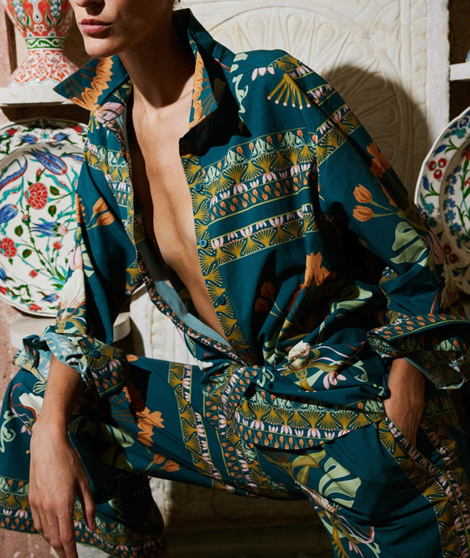 A person in Lug Von Siga's Franzie Top featuring a floral design leans against a tiled wall with decorative plates in the background.