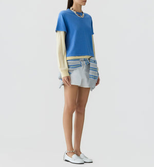 A woman wears the Guest in Residence Layer Tee, styled over cream cotton-silk undersleeves with a light mini skirt, striped sweater at her waist, white loafers, and a chunky necklace for an effortlessly layered look.