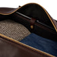 Unzip a Bennett Winch Leather Weekender from Bennett Winch, made with full grain leather, and reveal its contents: a folded tweed jacket, blue jeans, and a sleek black device.