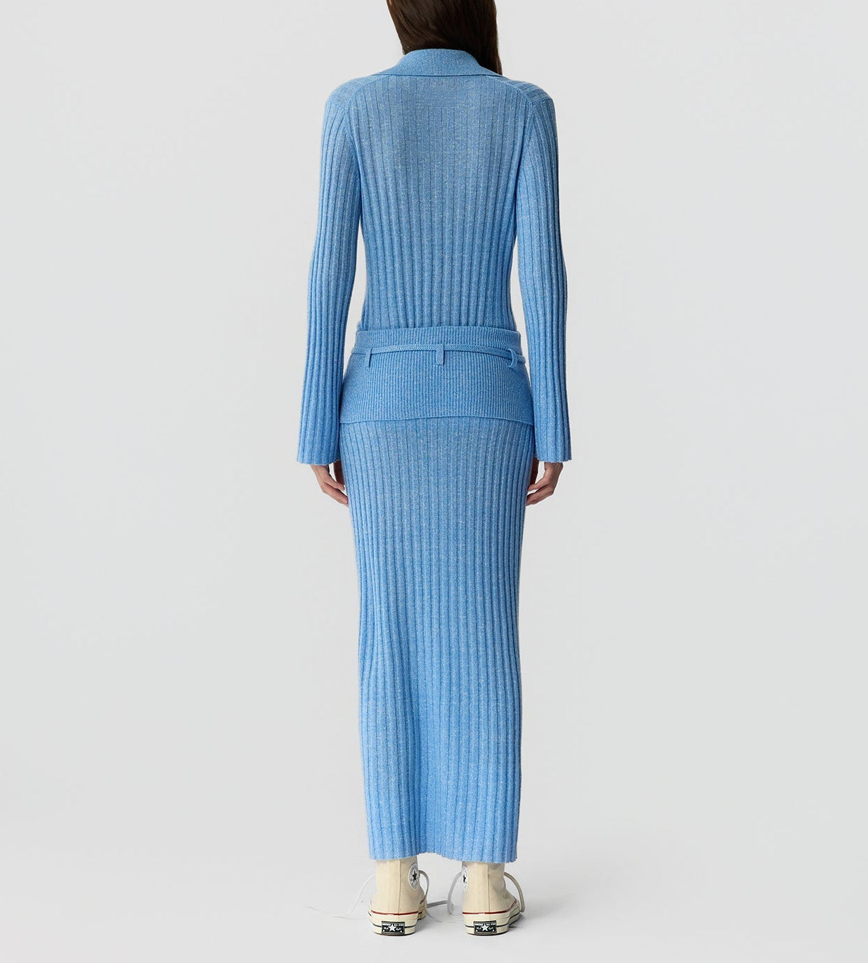 A person faces away, wearing the Lia Skirt by Guest in Residence—a long, ribbed, light blue knit skirt with maxi-inspired lines—paired with beige shoes against a plain light background.