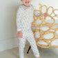 A young child stands barefoot on a light floor, wearing the Lila & Hayes Baby Boys' and Boys' Grayson Pajama Set, and holds onto a rattan chair in a bright room.