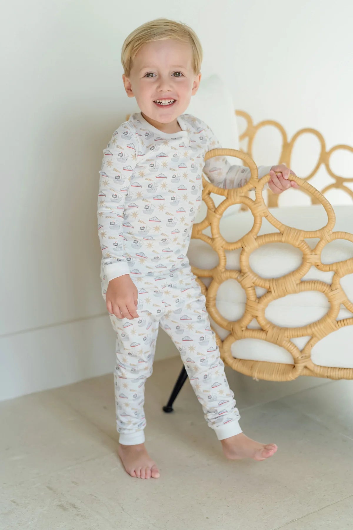 A young child stands barefoot on a light floor, wearing the Lila & Hayes Baby Boys' and Boys' Grayson Pajama Set, and holds onto a rattan chair in a bright room.