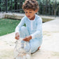 A young boy wears the Boys' Jack Pajama Set by Lila & Hayes, featuring light blue long sleeves and an elastic waistband, as he sits outdoors on a stone surface, placing seashells into a clear glass jar.