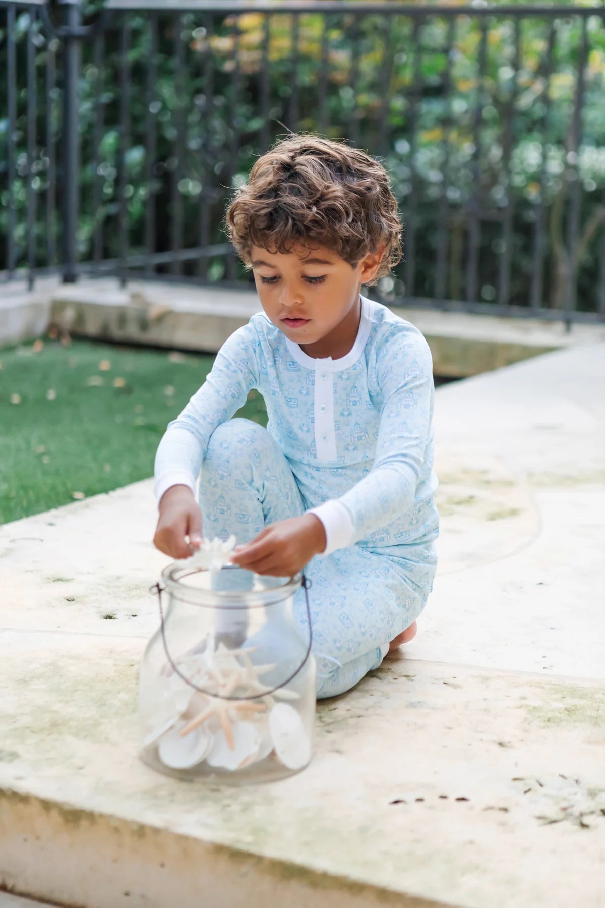 A young boy wears the Boys' Jack Pajama Set by Lila & Hayes, featuring light blue long sleeves and an elastic waistband, as he sits outdoors on a stone surface, placing seashells into a clear glass jar.