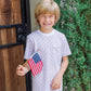 A smiling child in the Lila & Hayes Boys' Charles Short Set holds a small American flag while standing outside by a wooden door and green shrub.